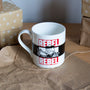 White mug with 'REBEL REBEL' design on a brown surface with wrapped gifts in the background