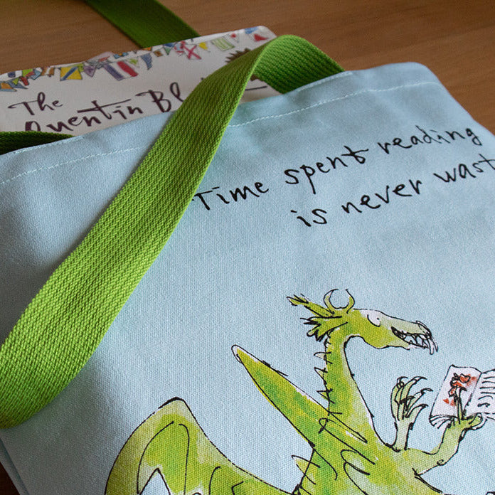 Tote bag with a dragon design and text, placed on a wooden surface with books underneath.