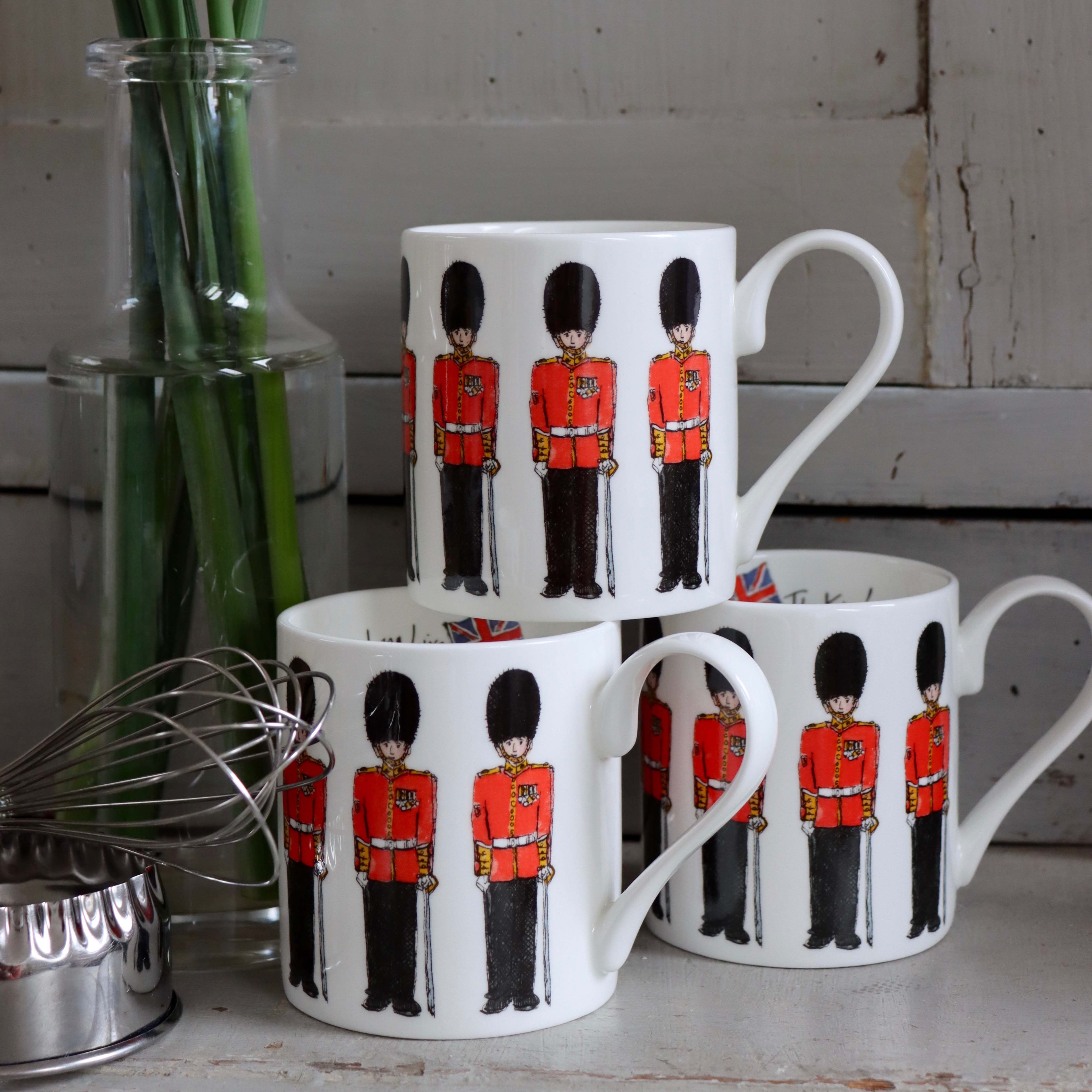 A set of white bone china mugs with illustrations of the King's Guard in red uniforms, displayed on a table with a flower vase and a sprinkle of sugar in the background.